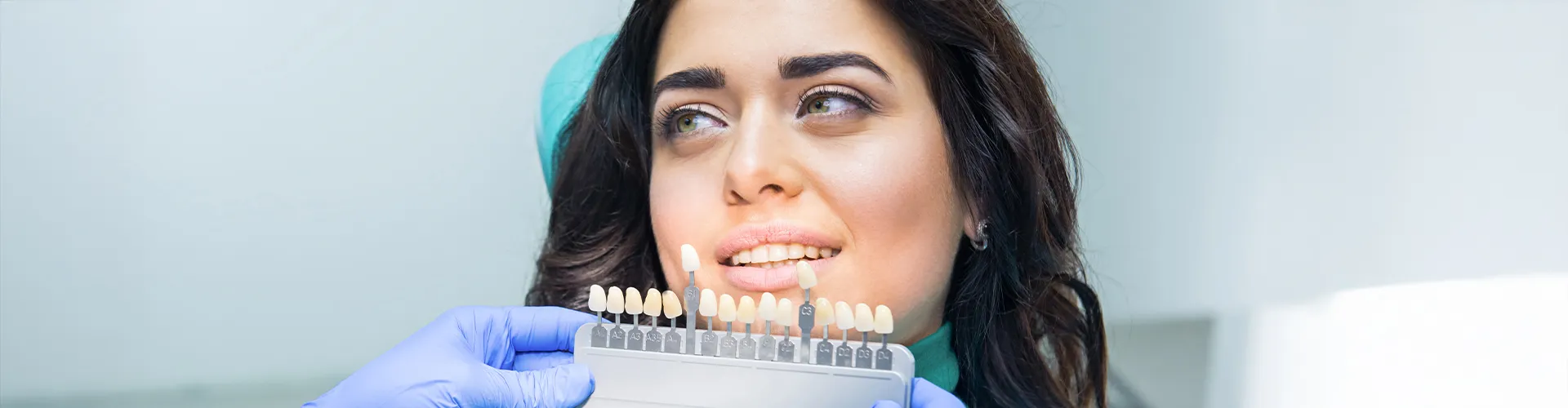 Gloved hand holding tooth shade-matching tool next to woman's smile.
