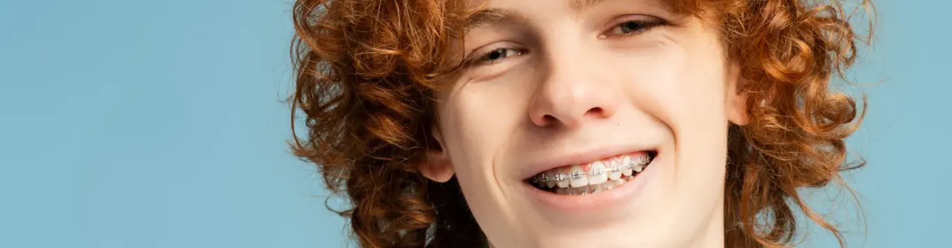 Smiling young patient wearing traditional metal braces against a light blue background.