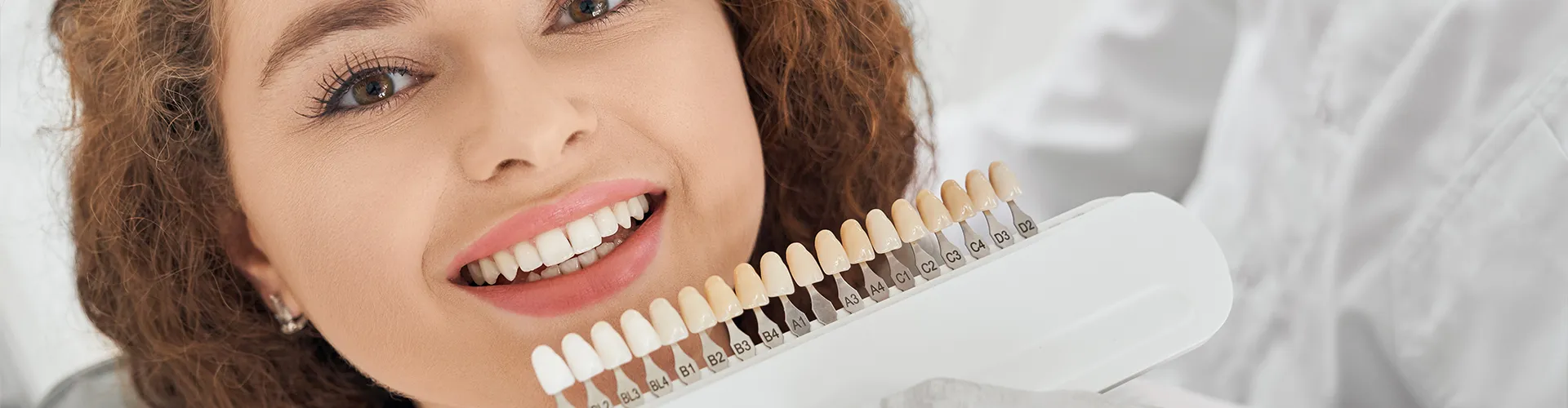 Gloved hand holding tooth shade-matching tool against woman's teeth.
