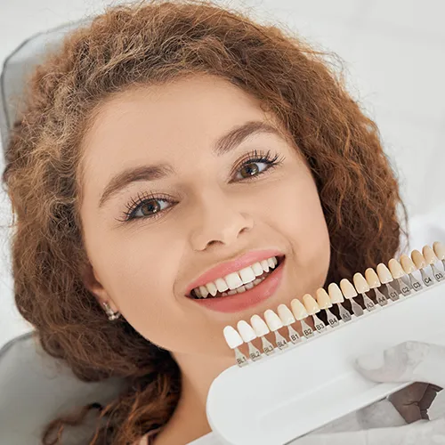 Gloved hand holding tooth shade-matching tool against woman's teeth.