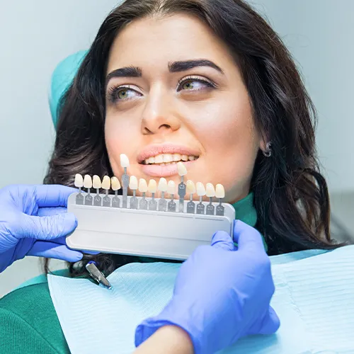 Gloved hand holding tooth shade-matching tool next to woman's smile.