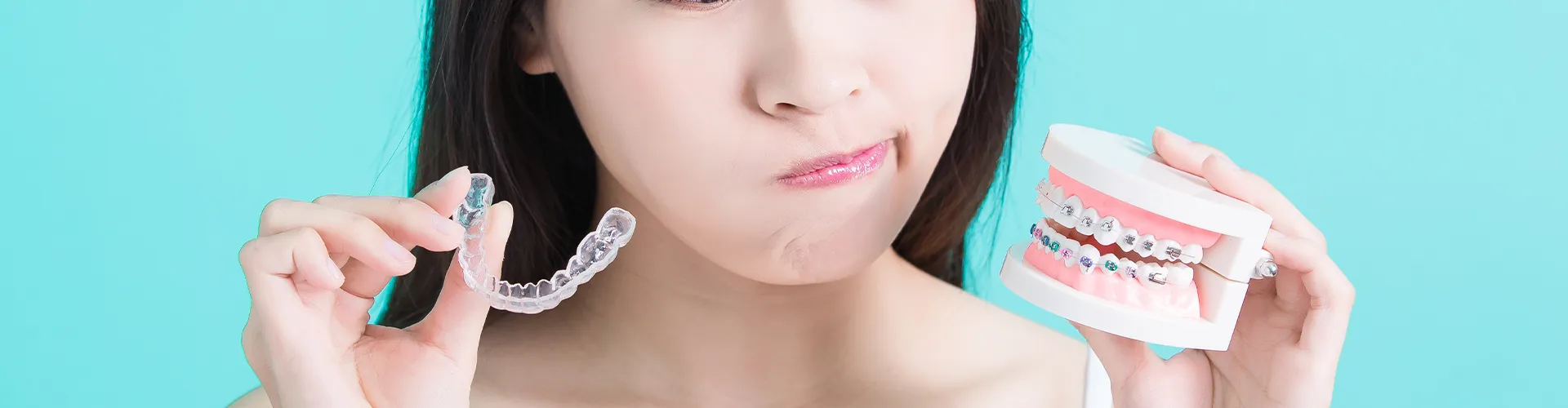 Woman holding clear aligner in one hand and mouth model wearing braces in the other.